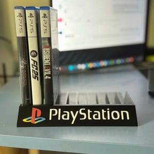 May include: A black plastic game storage stand featuring the PlayStation logo and text on the front. The stand holds three PS5 game cases, specifically Black Myth Wukong, FC25, and Resident Evil 4, in a vertical row within its designated slots.