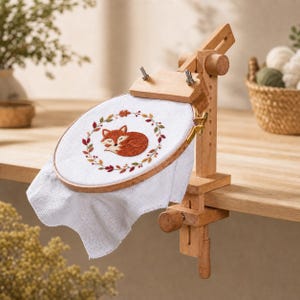 May include: A wooden embroidery hoop stand with an adjustable clamp attached to a table. The hoop holds white fabric featuring an embroidered sleeping orange fox surrounded by a circular wreath of small, colorful autumn-toned leaves.