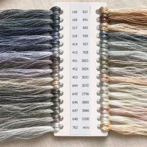 May include: A white plastic organizer card holding multiple strands of embroidery floss in various shades of gray, blue, and beige. The card features two columns of printed numerical color codes ranging from 168 to 5200 and the word ecru.