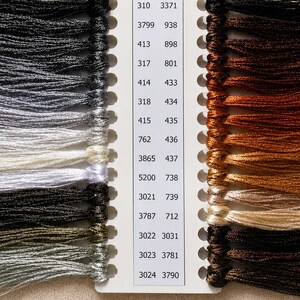 May include: A white thread organizer card featuring two columns of numerical color codes and corresponding embroidery floss samples. The left side displays shades of gray, black, and white, while the right side shows various brown and tan tones.