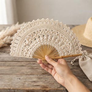 May include: A handheld folding fan featuring a cream-coloured crocheted lace cover with a scalloped edge and intricate openwork patterns, attached to a light brown bamboo frame with thin, radiating slats that extend from a central pivot point.