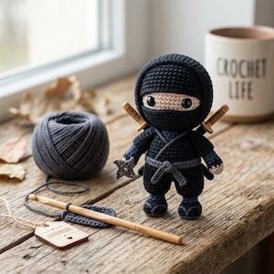 Op de afbeelding: Een gehaakte ninjapop in een zwart pak met een beige gezicht, zwarte ogen en twee zwaarden op de rug. Het houdt een grijze shuriken vast. Daarnaast liggen een bol donkergrijs garen, een houten 3,5 mm haaknaald en een label met "HANDMADE".