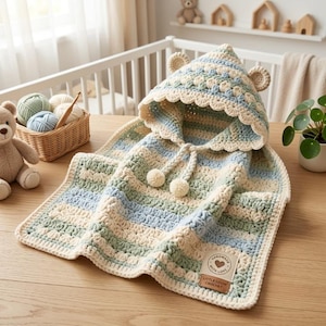 May include: A hooded crochet blanket featuring horizontal stripes in cream, sage green, and light blue. The hood has two small bear ears, a scalloped edge, and two pom-poms on drawstrings. A tag reads "Handmade with love" and "Little Loom Crochet".