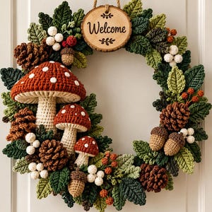 Op de afbeelding: Een ronde krans met gehaakte elementen, waaronder drie rood-witte paddenstoelen, bruine dennenappels, beige eikels, witte bessen en groene bladeren. Bovenaan hangt een rond houten bordje met de inscriptie Welcome.