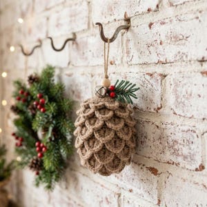 May include: A crocheted pinecone ornament in a light brown yarn, featuring a textured scale pattern. The ornament is topped with a small wooden bead, a silver jingle bell, two red berries, and a small green pine sprig, hanging from a jute twine loop.