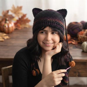 Puede incluir: Un gorro de ganchillo hecho a mano con orejas de gato en tonos oscuros variados de negro, marrón y morado intenso. El gorro presenta dos orejas puntiagudas en la parte superior y dos largas trenzas laterales, cada una terminada con una pequeña calabaza.