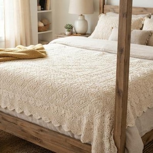 Könnte beinhalten: Eine cremefarbene gehäkelte Tagesdecke mit gewelltem Rand, die über einem hölzernen Himmelbett liegt. Eine senfgelbe Strickdecke ist am Fußende des Bettes gefaltet, das mit passenden cremefarbenen Kissen und Bettwäsche ausgestattet ist.