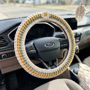 May include: A crochet steering wheel cover featuring horizontal stripes in cream, mustard yellow, and sage green, accented with a single three-dimensional cream-colored flower at the top. A matching cream-colored macrame feather hangs nearby.