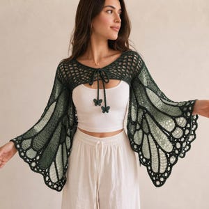 May include: A dark green crochet bolero featuring long, flared sleeves with a butterfly wing pattern. The garment has an open front with a tie closure adorned with two small dark green butterfly-shaped charms, worn over a white tank top.