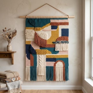 May include: A large rectangular wall hanging featuring a variety of textured crochet and knit patterns in teal, mustard yellow, cream, navy blue, and terracotta. The piece is suspended from a wooden dowel with a cord and has long fringe at the bottom.