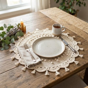 May include: A round, cream-colored crocheted placemat with a scalloped edge and tassel fringe. It features a speckled ceramic plate, a beige linen napkin, a silver fork, a silver knife, and a ceramic mug. A candle and eucalyptus are nearby.