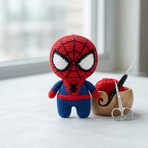 May include: A crocheted Spider-Man doll with a red head featuring black web patterns and large white eyes, a blue body with a red chest emblem, and a small black spider icon. Beside it sits a wooden yarn bowl with red yarn, a crochet hook, and scissors.