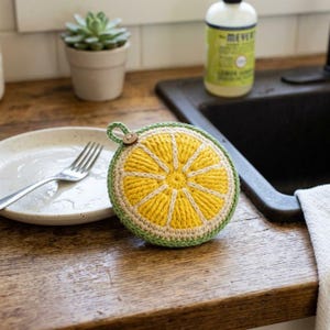 May include: A round, crocheted lemon slice scrubber featuring a bright yellow center with white segments, a light green border, and a small wooden button attached to a loop at the top. The design mimics the appearance of a citrus fruit slice.