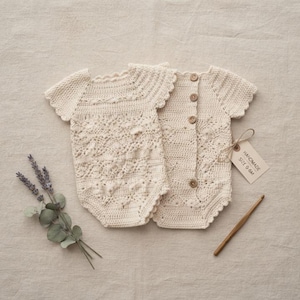 May include: Two cream-colored crochet rompers with short scalloped sleeves and intricate lace-like patterns. The rompers feature a vertical row of four small brown wooden buttons down the back. A tag attached with string reads HANDMADE SIZE 0-6M.