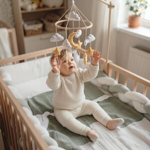 May include: A baby mobile featuring crocheted white clouds, yellow stars, and a yellow crescent moon suspended from a circular wooden frame. The mobile hangs above a wooden crib with a braided bumper and a soft green and white muslin blanket.