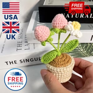 May include: A small crocheted potted plant featuring two round pink flower buds and one white flower with a yellow center. The plant has bright green leaves and stems, set in a tan crocheted pot with a textured brown top representing soil.