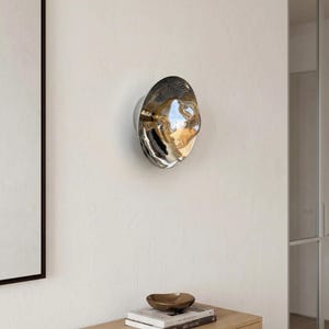 May include: A circular wall-mounted light fixture featuring a reflective, metallic, and amber-tinted glass surface with an irregular, organic, and undulating shape that creates a distorted mirror effect on the wall.