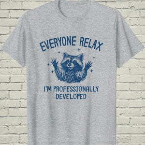 May include: A heather gray short-sleeve t-shirt featuring a dark blue graphic of a raccoon with its paws raised. The text reads EVERYONE RELAX I'M PROFESSIONALLY DEVELOPED in a bold blue font, surrounded by small star accents.