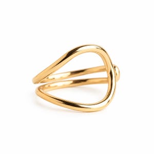 Könnte beinhalten: Ein polierter goldfarbener Ring mit einem offenen, fließenden und asymmetrischen wellenartigen Design. Das Band besteht aus zwei glatten, miteinander verbundenen Schlaufen, die eine durchgehende, organische Form mit glänzender Oberfläche bilden.