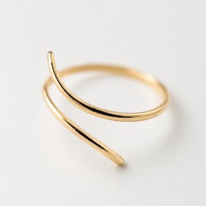 Puede incluir: Un anillo de tono dorado con un diseño de bypass que presenta una banda continua única que se envuelve para crear una forma de espiral abierta y superpuesta. La superficie metálica es lisa y pulida, reflejando la luz en la forma curva.