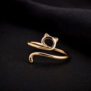 Può includere: Un anello aperto color oro che presenta la sagoma stilizzata di un gatto. La fascia si curva per formare il contorno di una testa di gatto con orecchie a punta sulla parte superiore e una coda sottile che si estende lungo la parte inferiore.