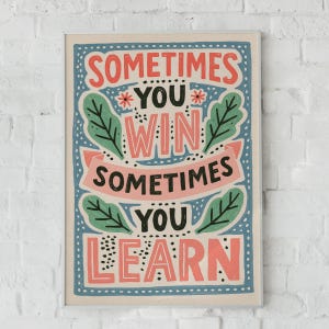 May include: A rectangular wall art print featuring the text "SOMETIMES YOU WIN SOMETIMES YOU LEARN" in a mix of black and coral pink lettering. The design includes green leaf illustrations, small red flowers, and a coral pink banner on a blue background.