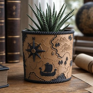 May include: A cylindrical planter with a tan surface featuring a raised black map design, including a compass rose, a sailing ship, and mountain ranges. The rim and base are finished with a dark rope-like texture. A green succulent plant sits inside.