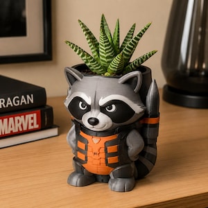 May include: A 3D-printed planter shaped like a raccoon character wearing an orange and black tactical vest. The raccoon has gray fur, a black eye mask, and a striped tail. A green succulent plant with white horizontal stripes sits inside the planter.