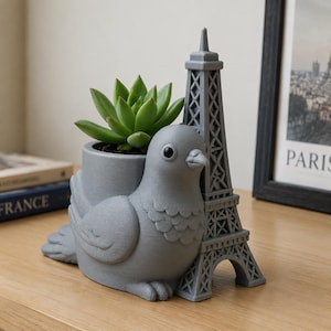 May include: A gray 3D-printed planter shaped like a pigeon sitting next to a miniature Eiffel Tower. The pigeon has a cylindrical opening on its back containing a green succulent plant. The structure is matte gray with visible layer lines.