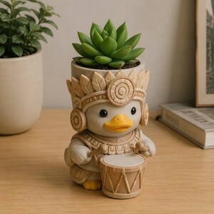Puede incluir: Una maceta beige con forma de pato que lleva un tocado de plumas y una pechera ornamentados, sostiene dos baquetas y está de pie detrás de un tambor. Una planta suculenta verde está plantada en la parte superior del recipiente.