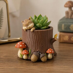 May include: A brown tree stump planter featuring a sleeping chipmunk figurine on the rim, a green succulent plant, two red and white spotted mushrooms, and two brown acorns attached to the base, with a small branch extending from the side.