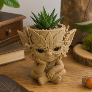 May include: A light brown, sculpted planter shaped like a forest creature with large eyes, pointed ears, and a face framed by carved leaves. The creature sits with one hand resting on its chin, holding a small, spiky green succulent plant.