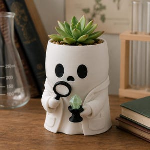 Pode incluir: Um vaso branco em forma de fantasma vestindo um jaleco, segurando uma lupa preta e um pequeno cristal verde. O fantasma tem olhos pretos grandes e uma boca pequena. Uma planta suculenta verde está plantada no topo da figura do fantasma.