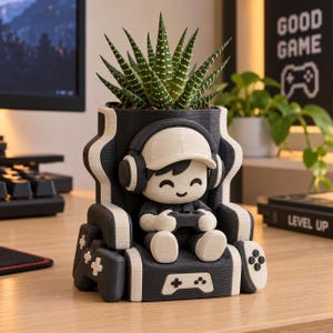 May include: A black and white 3D printed planter shaped like a gaming chair with a character wearing headphones and holding a controller. The planter holds a green succulent plant. The base features a game controller icon and side accents.