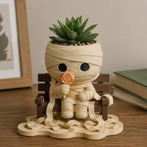 Puede incluir: Una maceta beige con forma de momia que contiene una planta suculenta verde. La figura de la momia está sentada en un banco de madera marrón y sostiene una pequeña piruleta de color naranja y blanco. Está envuelta en vendas que cuelgan.