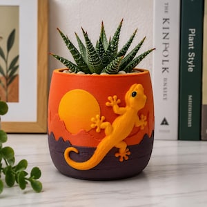 Puede incluir: Una maceta cilíndrica de color naranja y morado oscuro con un relieve de un gecko amarillo y un diseño de sol. La maceta contiene una planta suculenta verde y puntiaguda con patrones blancos moteados en sus hojas, evocando un paisaje desértico.