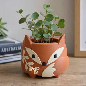 Puede incluir: Una maceta de cerámica color terracota con forma de cabeza de zorro, con orejas puntiagudas, ojos negros y nariz negra. La maceta presenta detalles decorativos blancos, incluyendo un diseño de rama con hojas en el lateral y una cola blanca en la base.