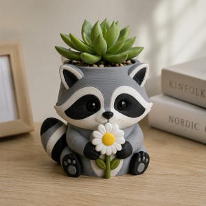 May include: A gray and white raccoon-shaped planter holding a white daisy with a yellow center. The raccoon has black eye patches, a striped tail, and small paws. A green succulent plant is potted inside the top opening of the raccoon figure.