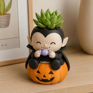 May include: A succulent plant sits in a planter shaped like a vampire character wearing a black cape and holding a small purple candy. The vampire is perched atop an orange jack-o'-lantern pumpkin with a carved face, featuring black eyes and a mouth.