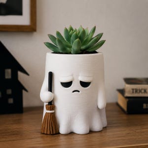 Pode incluir: Um vaso de cerâmica branca em forma de fantasma triste segurando uma vassoura castanha, contendo uma planta suculenta verde. A figura do fantasma tem olhos caídos e uma pequena boca voltada para baixo, com uma superfície texturizada.