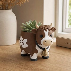 May include: A brown and white cow-shaped planter featuring a green succulent plant. The planter has a black collar with a gold-coloured bell charm, a white flower pattern on the side, and four black hooves. The cow has a tan muzzle and horns.
