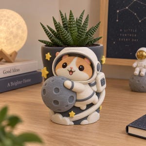 以下が含まれることがあります： 白い宇宙服とヘルメットを着用し、質感のある灰色の月を抱えた宇宙飛行士のハムスターが彫刻された、濃い灰色の円筒形の植木鉢。デザインには小さな黄色い星と白い軌跡が含まれており、緑色の多肉植物が植えられています。