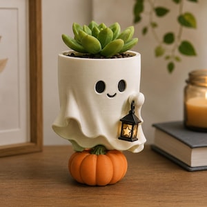 May include: A white ceramic planter shaped like a ghost with a smiling face, holding a small black lantern with a star cutout. The ghost sits atop a round orange pumpkin, and a green succulent plant is potted inside the top of the ghost figure.