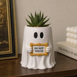 Pode incluir: Um vaso branco em forma de fantasma com dois olhos ovais pretos, segurando uma moldura retangular dourada que contém o texto preto DO NOT TOUCH. Uma planta suculenta verde está plantada na abertura superior da figura do fantasma.