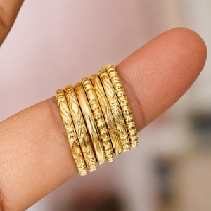 May include: A set of seven gold-toned rings stacked together, each featuring a distinct texture or pattern. The collection includes bands with beaded, smooth, and engraved geometric designs, all displayed in a uniform metallic finish.