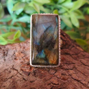 May include: A rectangular labradorite gemstone ring set in a silver-toned metal frame. The stone displays iridescent flashes of blue, gold, and dark gray across its polished surface, held by a beaded metal border surrounding the rectangular setting.