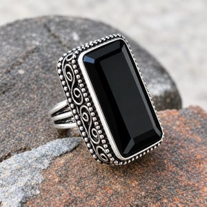 May include: A silver ring featuring a large, rectangular, faceted black stone set in a frame with beaded edges and intricate scrollwork patterns along the sides. The metal band has a polished finish and a multi-layered design on the shank.