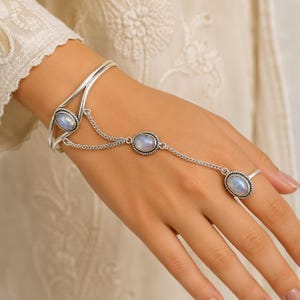 Op de afbeelding: Een zilveren slavenarmband met drie ovale, iriserende maanstenen in gedraaide metalen zettingen. Het sieraad bestaat uit een scharnierende armband die via fijne zilveren kettinkjes verbonden is met een centrale steen en een bijpassende ring.