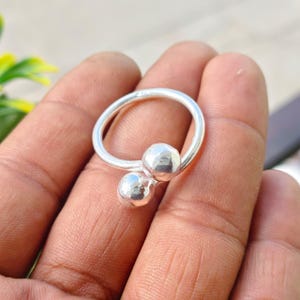 以下が含まれることがあります： 両端に磨かれた球状の金属ボールが付いたオープンバンドデザインの銀色の指輪。指輪は滑らかで光沢のある表面を持ち、円形をしています。2つの球体は開口部で互いに近くに配置されています。