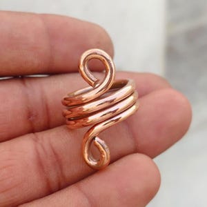 Puede incluir: Un anillo de cobre con un diseño de alambre continuo que se enrolla varias veces alrededor del dedo. El alambre termina en dos bucles en espiral redondeados y distintos en la parte superior e inferior, creando un acabado pulido y brillante.